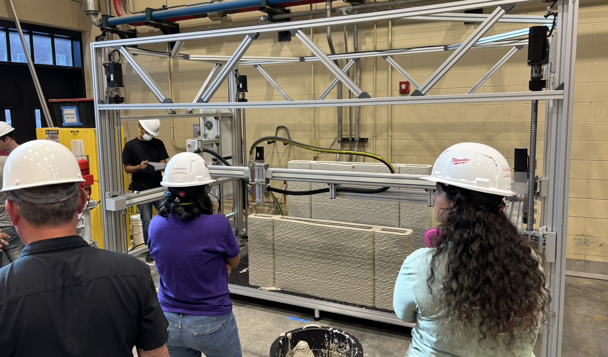 University students in hard hats watching the Build Additive gantry print a wall in a lab